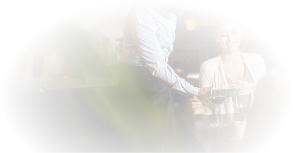 Kelner podaje danie uśmiechniętemu gościowi w stylowej restauracji, na stole stoją kieliszki do wina, a atmosfera jest ciepła i przyjazna.